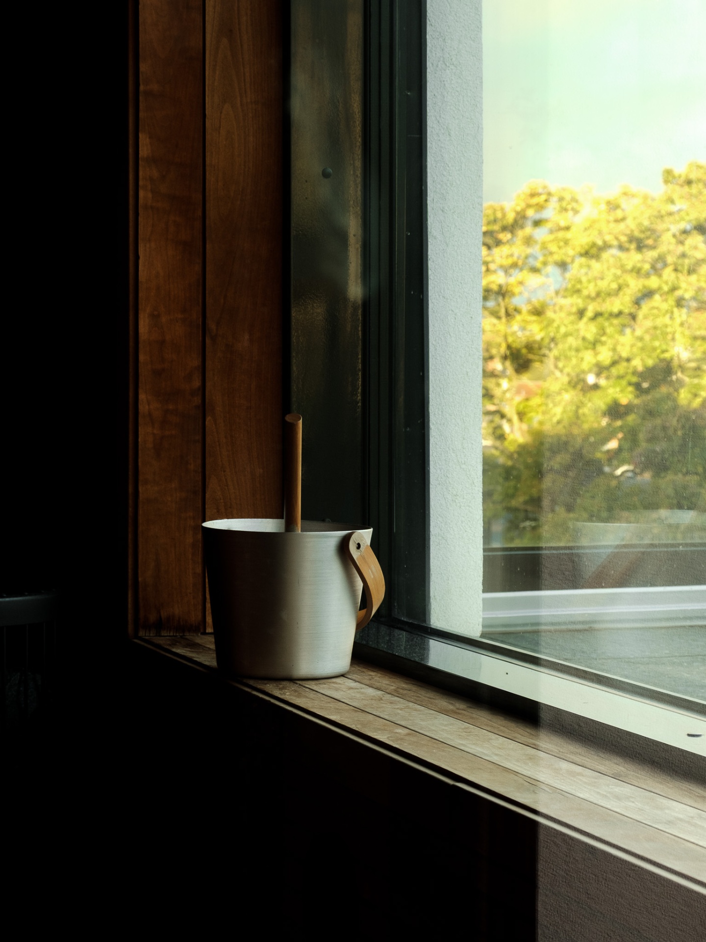 Sauna bucket and ladle on a wooden sill with sunlit foliage outside