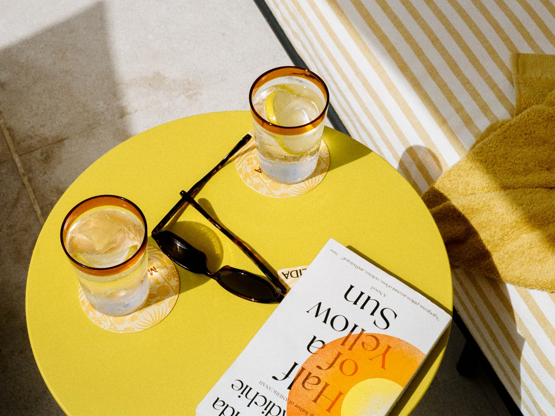 Yellow side table with iced water, sunglasses and a novel by the pool