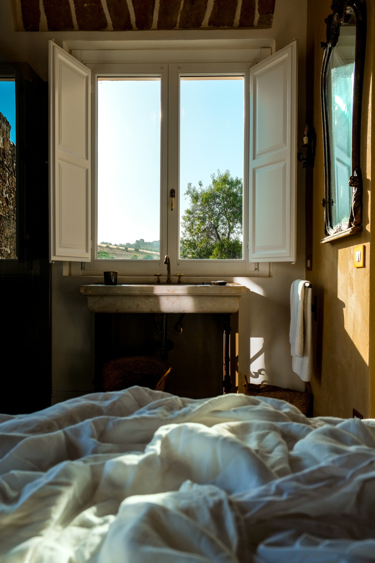 Unmade bed framing a hillside view through an open shuttered window
