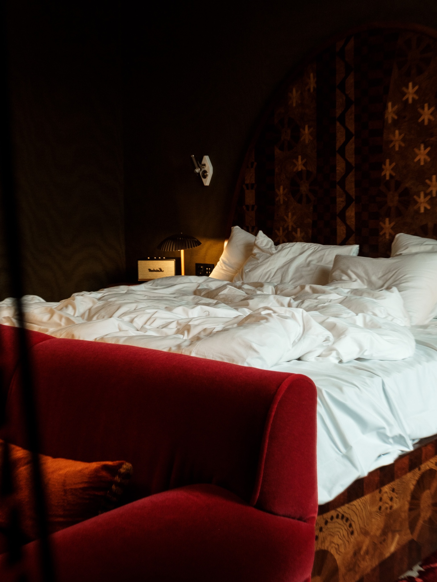 Unmade bed with red velvet armchair and patterned marquetry headboard