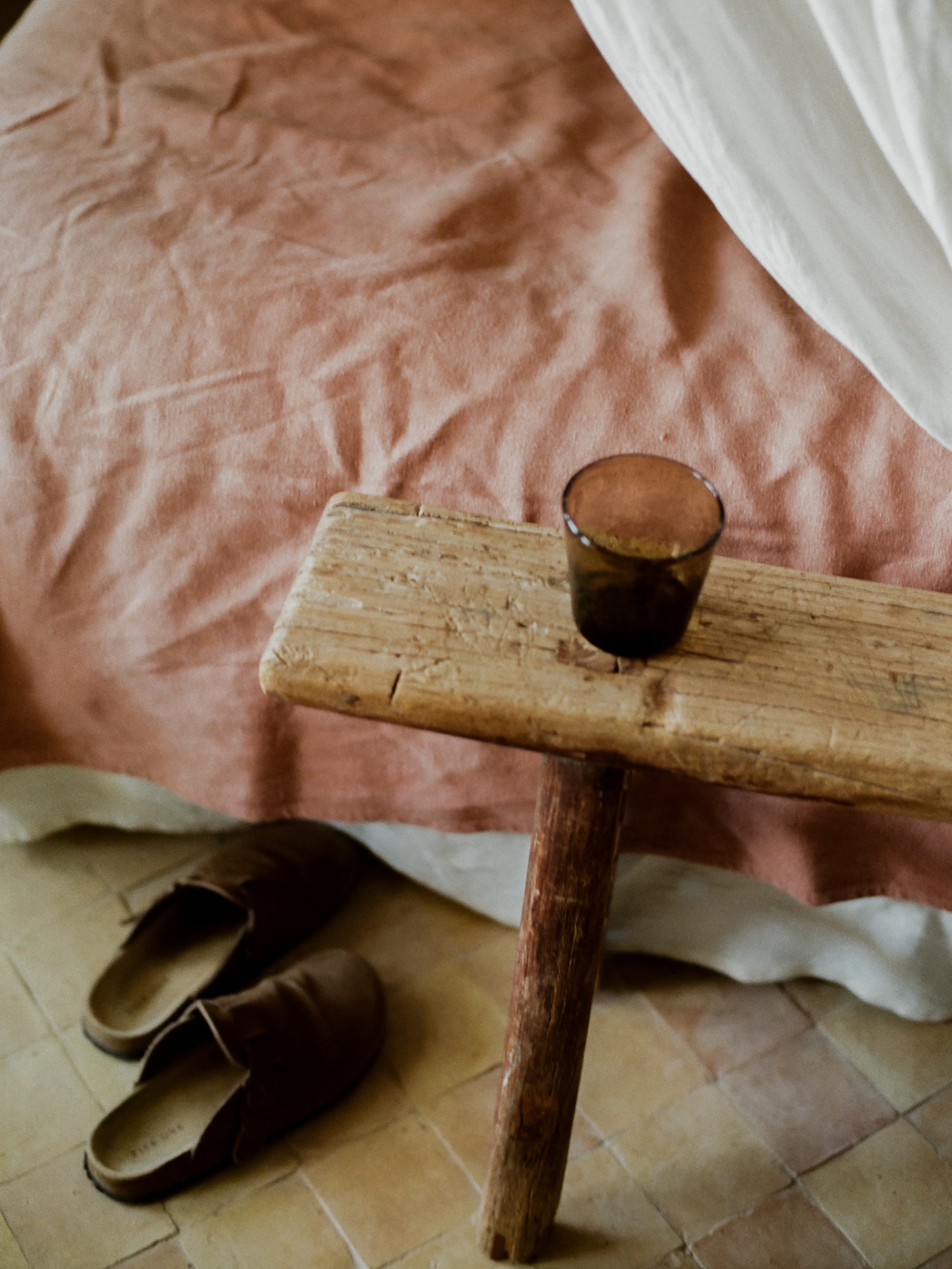 Wooden stool with amber glass beside dusty pink linen and clogs